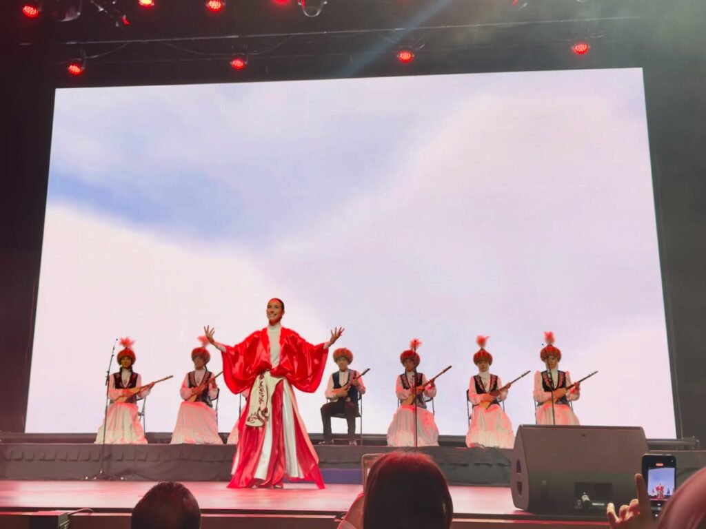 A young performer in traditional red costume singing on stage with musicians at the 24th IFLC Symphony of the Earth